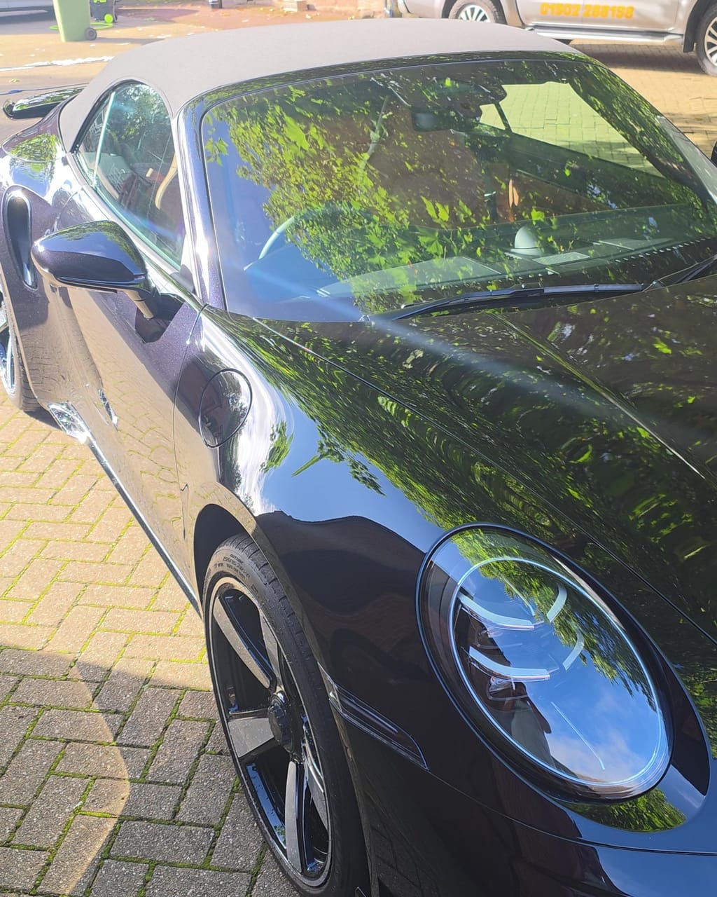 The same black Porsche 911 Turbo S after its fortnightly maintenance valet, headlight and wing reflecting the trees overhead